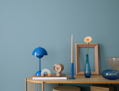 Blau-türkisfarbene Wand vor der ein kleiner Holztisch steht, auf dem Dekoelemente wie Vasen, Bilderrahmen und eine Tischlampe in Blautönen dekoriert sind